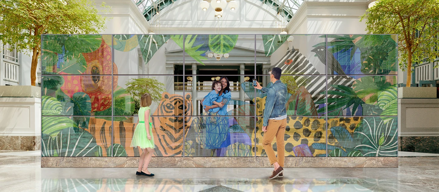 A family is taking a photo with a transparent OLED wall made up of 3ⅹ5 55EW5P indoors. The father, standing in front of the wall, is taking a picture of the mother and child, who are standing behind the wall with animals and plants naturally displayed on the screen.